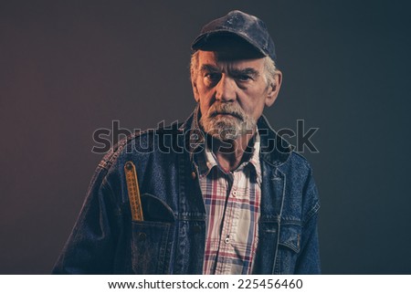 Senior carpenter with gray hair and beard holding yellow measuring rod wearing blue cap with jeans jacket. Low key studio shot.
