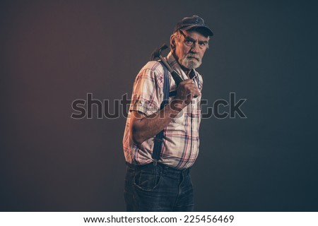 Senior male carpenter with gray hair and beard holding a hammer wearing blue cap with braces and jeans. Low key studio shot.