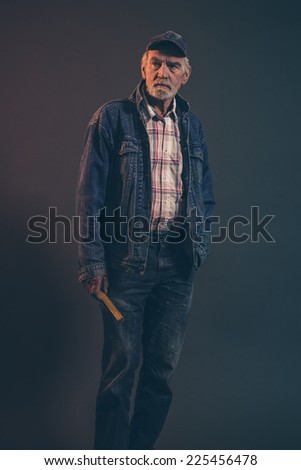Senior carpenter with gray hair and beard holding yellow measuring rod wearing blue cap with jeans jacket. Low key studio shot.