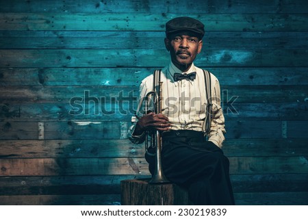 Vintage african american senior jazz musician with trumpet in front of old wooden wall. Wearing suit and cap.