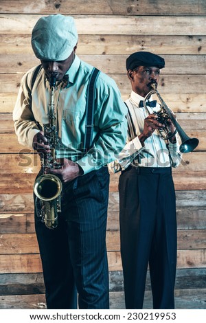 Two african american jazz musicians playing trumpet and saxophone. Standing in front of old wooden wall.