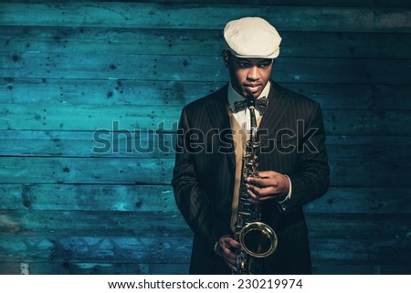 Vintage african american jazz musician with saxophone in front of old wooden wall. Wearing suit and cap.