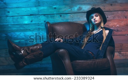 Cowgirl Wearing blue jeans and brown hat. Sitting on leather chair. In front of wooden wall.