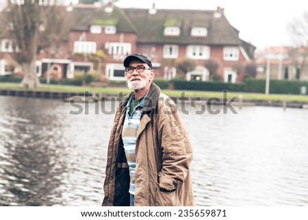 Senior man with beard wearing glasses and black cap outdoor in winter landscape with houses in the background.