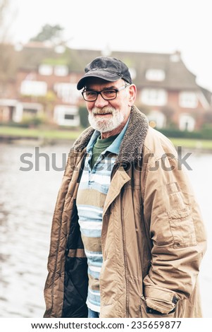 Senior man with beard wearing glasses and black cap outdoor in winter landscape with houses in the background.