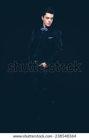 Retro fashion man with blue shirt, jacket, jeans and bow tie. Short dark hair. Studio shot against black.
