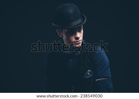 Vintage fashion man with blue shirt, gilet, jeans and bow tie. Wearing black hat. Studio shot against black.