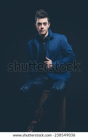 Winter jeans fashion man with short dark hair. Wearing blue jeans and jacket. Sitting on old wooden box. Studio shot against black.