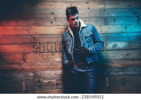 Winter jeans fashion man with short dark hair. Wearing jeans jacket, trousers and brown leather gloves. Leaning against old wooden wall.