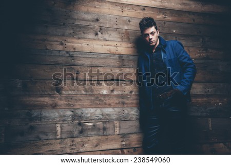 Winter jeans fashion man with short dark hair. Wearing blue jeans, jacket and brown leather gloves. Leaning against old wooden wall.