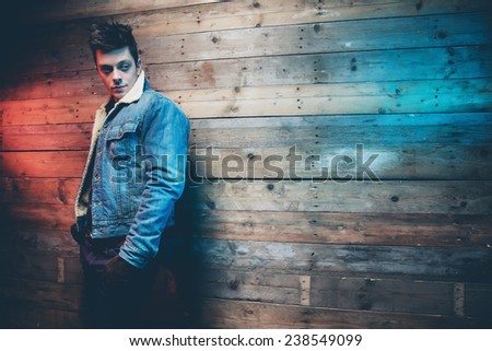 Winter jeans fashion man with short dark hair. Wearing jeans jacket, trousers and brown leather gloves. Leaning against old wooden wall.