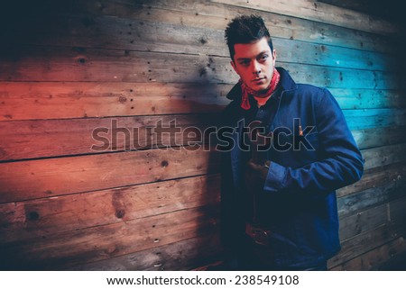 Winter jeans workwear fashion man with short dark hair. Holding old big spanner. Leaning against old wooden wall.