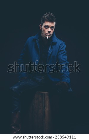 Winter jeans fashion man with short dark hair smoking a cigarette. Wearing blue jeans, jacket, brown leather boots and gloves. Sitting on old wooden box. Studio shot against black.