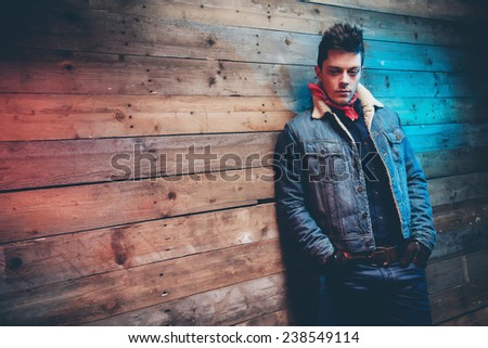 Winter jeans fashion man with short dark hair. Wearing jeans jacket and trousers. Leaning against old wooden wall.