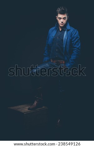 Winter jeans fashion man with short dark hair. Wearing blue jeans, jacket, brown leather boots and gloves. Standing on old wooden box. Studio shot against black.