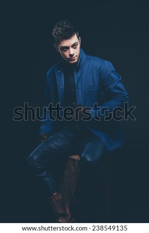 Winter jeans fashion man with short dark hair. Wearing blue jeans, jacket, brown leather boots and gloves. Sitting on old wooden box. Studio shot against black.