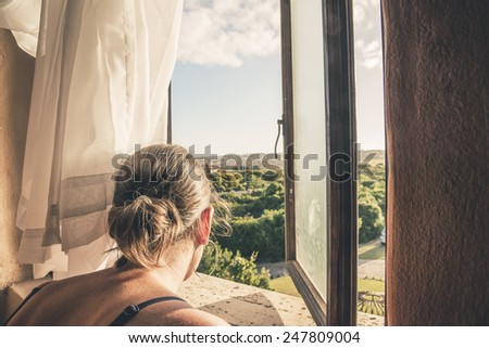 Over the shoulder shot of brunette woman looking through window at green summer landscape.