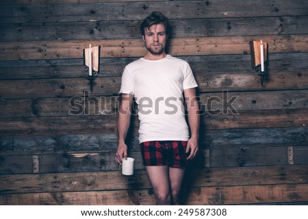 Man with blonde hair wearing white t-shirt and red flannel shorts. Standing against wooden wall inside wooden cabin.