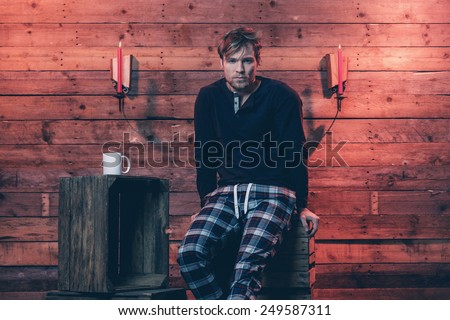 Man with blonde hair and beard wearing winter sleepwear. Sitting on wooden crate inside cabin.