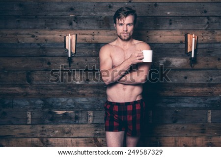 Man with blonde hair and bare chest wearing red flannel shorts. Standing against wooden wall inside wooden cabin.