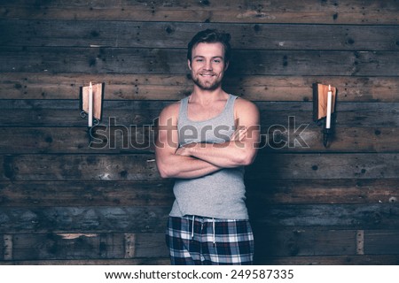Man with blonde hair wearing gray singlet shirt and blue flannel pants. Standing against wooden wall inside wooden cabin.