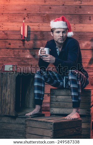 Man with blonde hair, beard and christmas hat wearing winter sleepwear. Sitting on wooden box inside cabin.