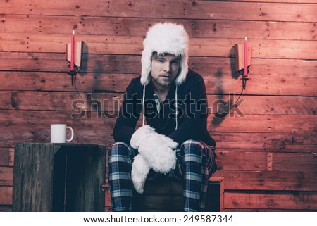 Man with blonde hair, beard and white hat wearing winter sleepwear. Sitting on wooden box inside cabin.
