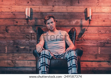 Man with blonde hair wearing winter sleepwear. Sitting in leather chair inside wooden cabin.