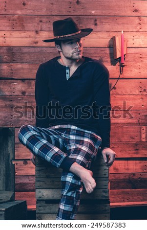 Man with blonde hair and brown cowboy hat wearing winter sleepwear. Sitting on wooden box inside cabin.
