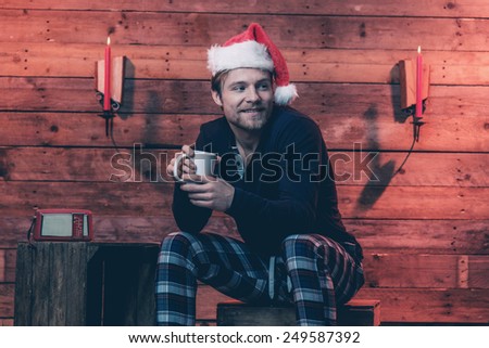 Man with blonde hair, beard and christmas hat wearing winter sleepwear. Sitting on wooden box inside cabin.