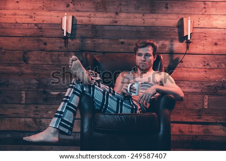Man with blonde hair wearing winter sleepwear. Sitting in leather chair inside wooden cabin.