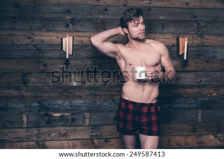 Man with blonde hair and bare chest wearing red flannel shorts. Standing against wooden wall inside wooden cabin.