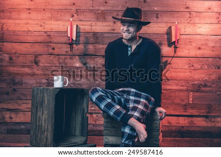 Man with blonde hair and brown cowboy hat wearing winter sleepwear. Sitting on wooden box inside cabin.
