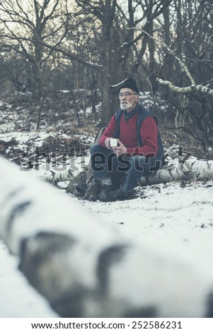 Active senior man outdoors in winter woodland sitting in his warm winter clothing on a fallen tree trunk in the snow enjoying a mug of hot coffee