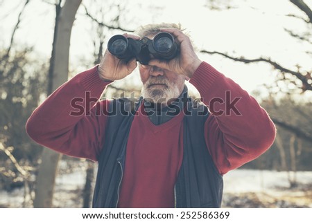 Elderly bearded man birdwatching in winter standing outdoors in a snowy forest scanning his surroundings with binoculars