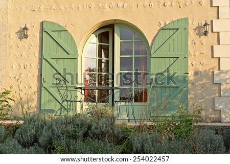 Arched glass front door with green shutters standing slightly ajar in evening sunshine in a beige textured exterior wall of a house