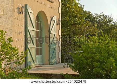 Arched entrance door to a house with shutters in a rough textured rustic exterior wall surrounded by greenery in evening sunlight