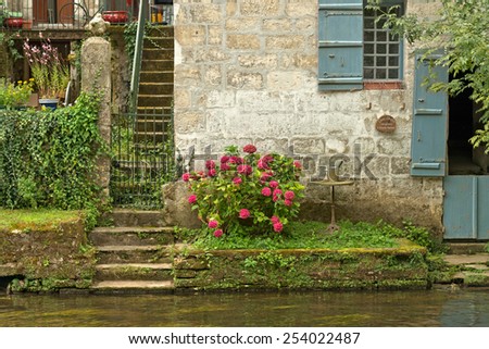Steps leading down to a river or canal from the door of an old cottage with blue shutters and trailing plants over stone walls