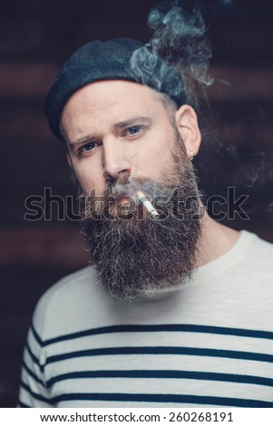Close up Gorgeous Man with Long Goatee, Wearing a Trendy Shirt and a Cap, Smoking a Cigarette While Looking at the Camera.