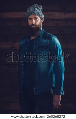 Serious Young Man with Long Beard Wearing Casual Denim Jacket and Knitted Hat Outfit, Standing In Front a Wooden Wall Background.