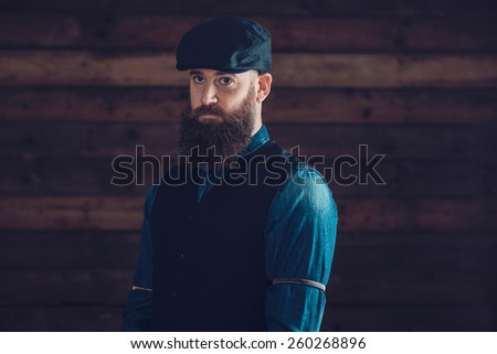 Half Body Shot of a Middle Age Man with Long Goatee Wearing a Trendy Attire and an Ivy Hat on a Wooden Wall Background.