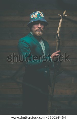 Portrait of a Serious Middle Age Man with Goatee in Green Formal Wear with hat Holding a Dry Tree Branch in front of a Wooden Wall While Looking at the Camera