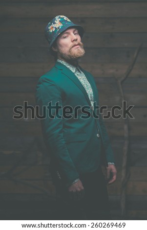 Portrait of a Middle Age Man with Goatee Wearing Elegant Green Formal Outfit and Hat, Standing in Front of a Wooden Background with Horizontal orientation While Looking at the Camera.