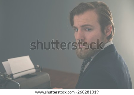 Handsome bearded vintage businessman sitting at his desk with an old manual typewriter turning to look thoughtfully at the camera, aged effect