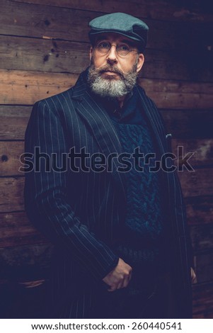 Portrait of a Serious Adult Goatee Man in Black Formal Outfit with Ivy Cap Standing In Front a Wooden Wall While Looking at the Camera.
