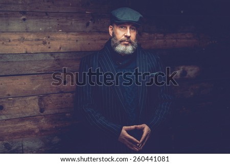 Half Body Shot of a Senior Goatee Man Wearing Black Formal Outfit with Ivy Cap, Posing in Front a Wooden Wall with his Hands In Front his Belly While Looking at the Camera.