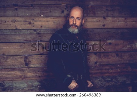 Adult Bald Goatee Man in Black Trendy Outfit Posing In front a Wooden Wall Background While Looking at the Camera.