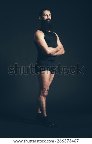 Portrait of a Muscular Man with Long Facial Hair Standing Confidently with Arms and Legs Crossed and Looking at the Camera. Captured in Studio with Black Background.
