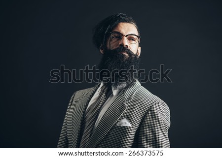 Thoughtful Man with Long Facial Hair Wearing Checkered Formal Wear with Glasses, Looking to the Right of the Frame Seriously on a Black Background.