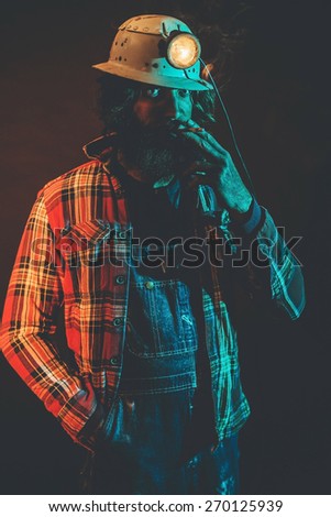 Male Miner Wearing Lit Safety Helmet Lamp, Overalls and Plaid Shirt Standing with Hand in Pocket and Smoking Cigarette in Studio with Blue Light